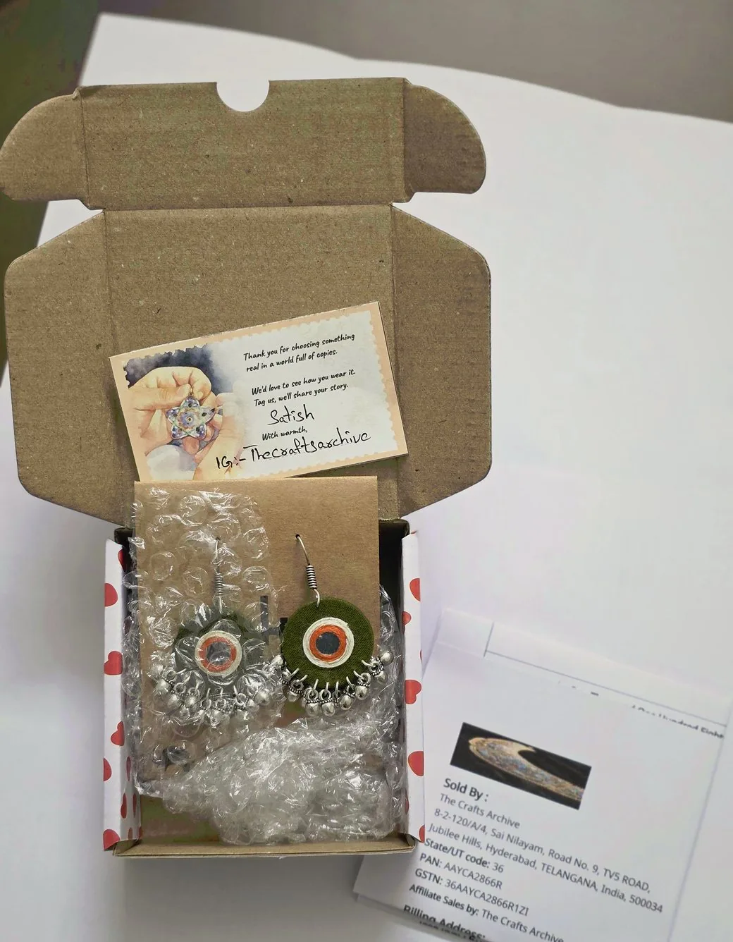 A real Wcommerce unboxing: plain brown outer box, handwritten thank-you card signed by Satish of The Crafts Archive, a pair of handmade earrings nested in heart-print tissue and bubble wrap, and a seller-branded invoice.